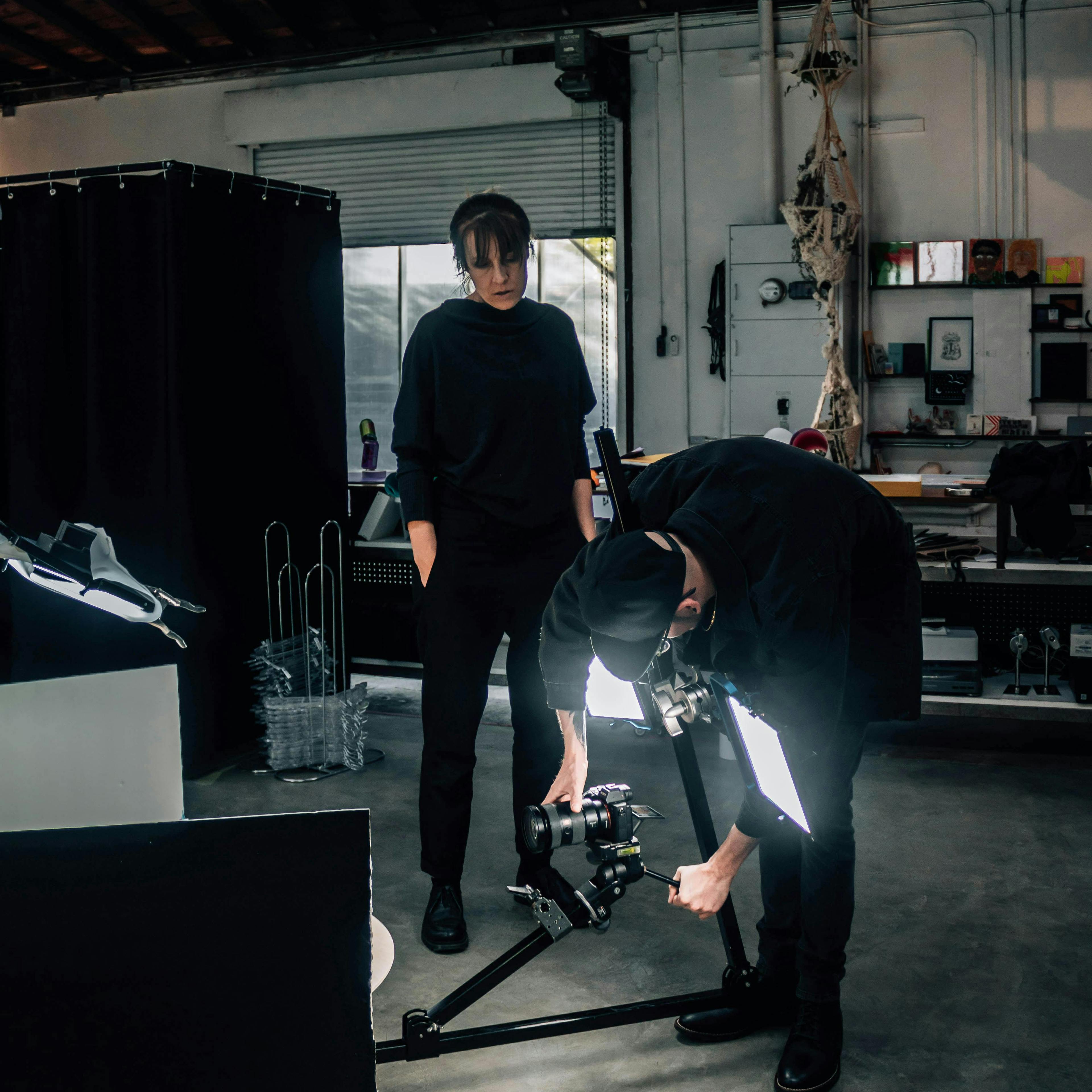 Two photographers working together on a 360 rig in a photography studio. One photographer is adjusting the rig while the other is framing the shot. The studio is well-lit and filled with photography equipment, including lights, reflectors, and cameras.