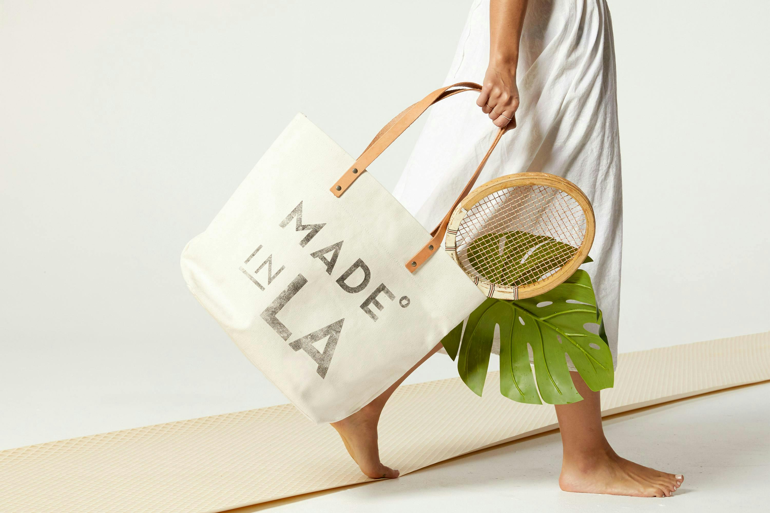 canvas tote bag for Cisco's company made in LA  photographed with a racket and monstera leave being held by a woman in a long dress barefoot 