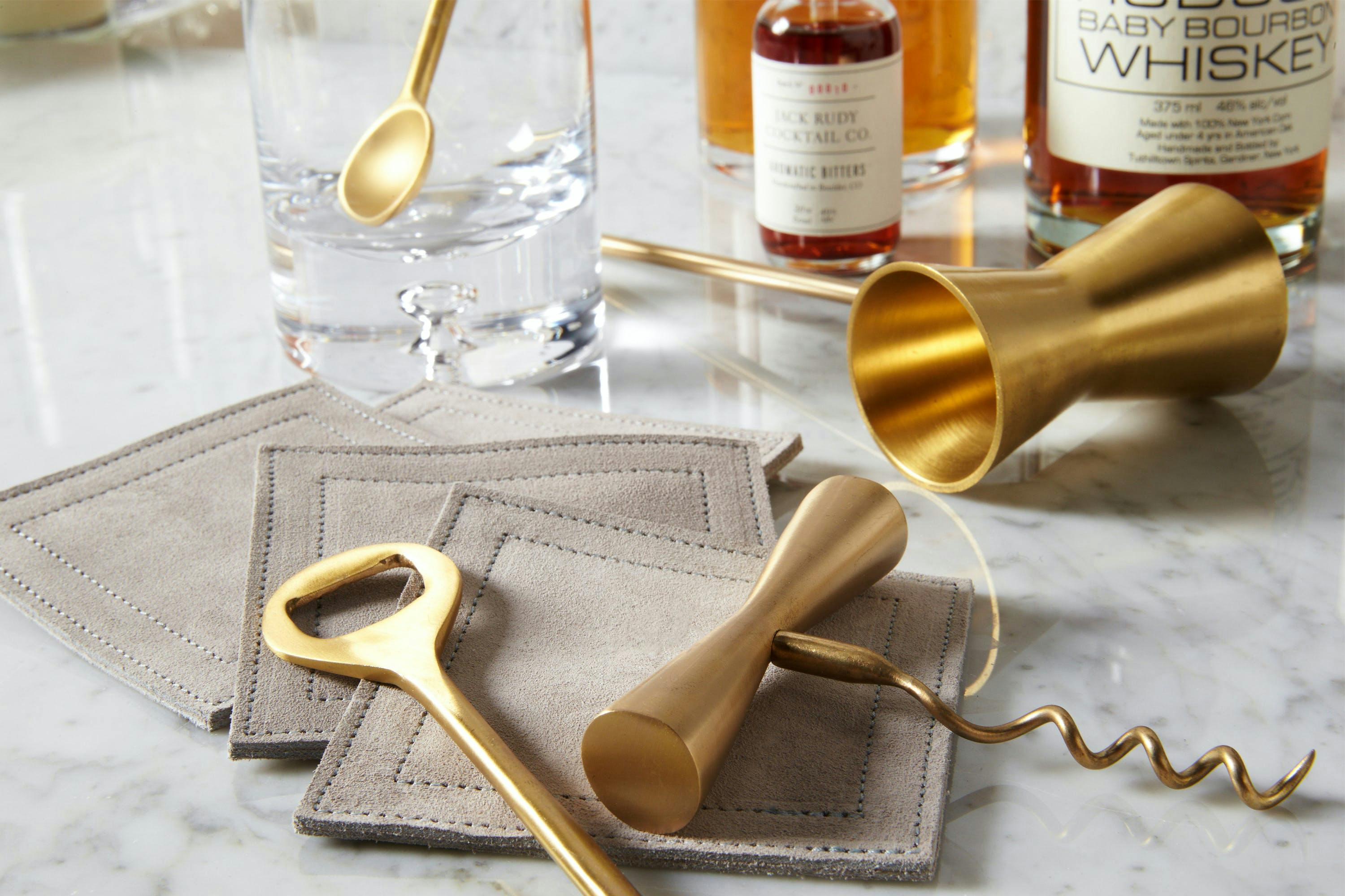 Flat lay of alcohol accessories on a marble table. Items include a cocktail shaker, jigger, mixing spoon, strainer, and citrus peeler. The marble table serves as the background