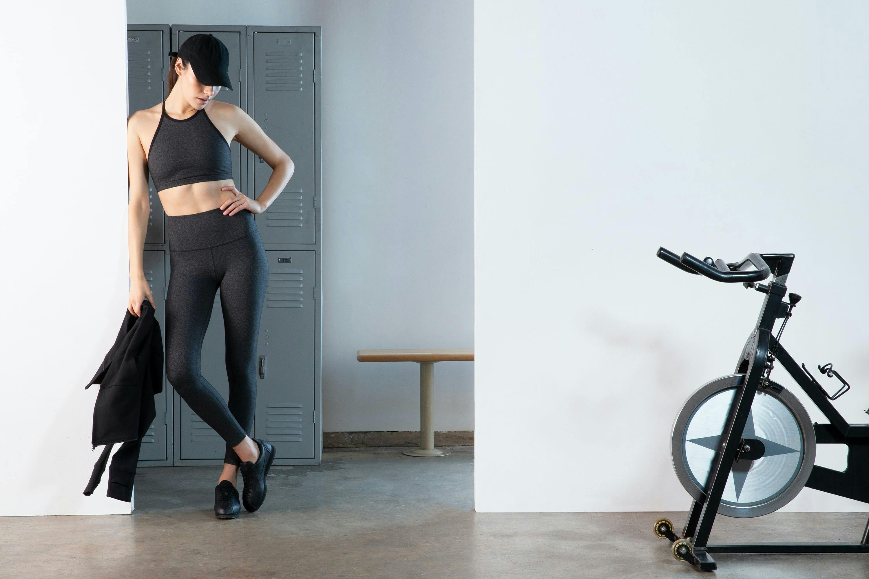 fashion photography of a woman posing against a wall in beyond yoga clothes with hand on hip in a locker room with a stationary bike on the right 